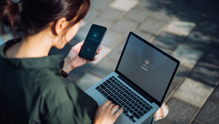 Young Asian businesswoman sitting on the bench in an urban park working outdoors, logging in to her laptop and holding smartphone on hand with a security key lock icon on the screen. Privacy protection, internet and mobile security concept