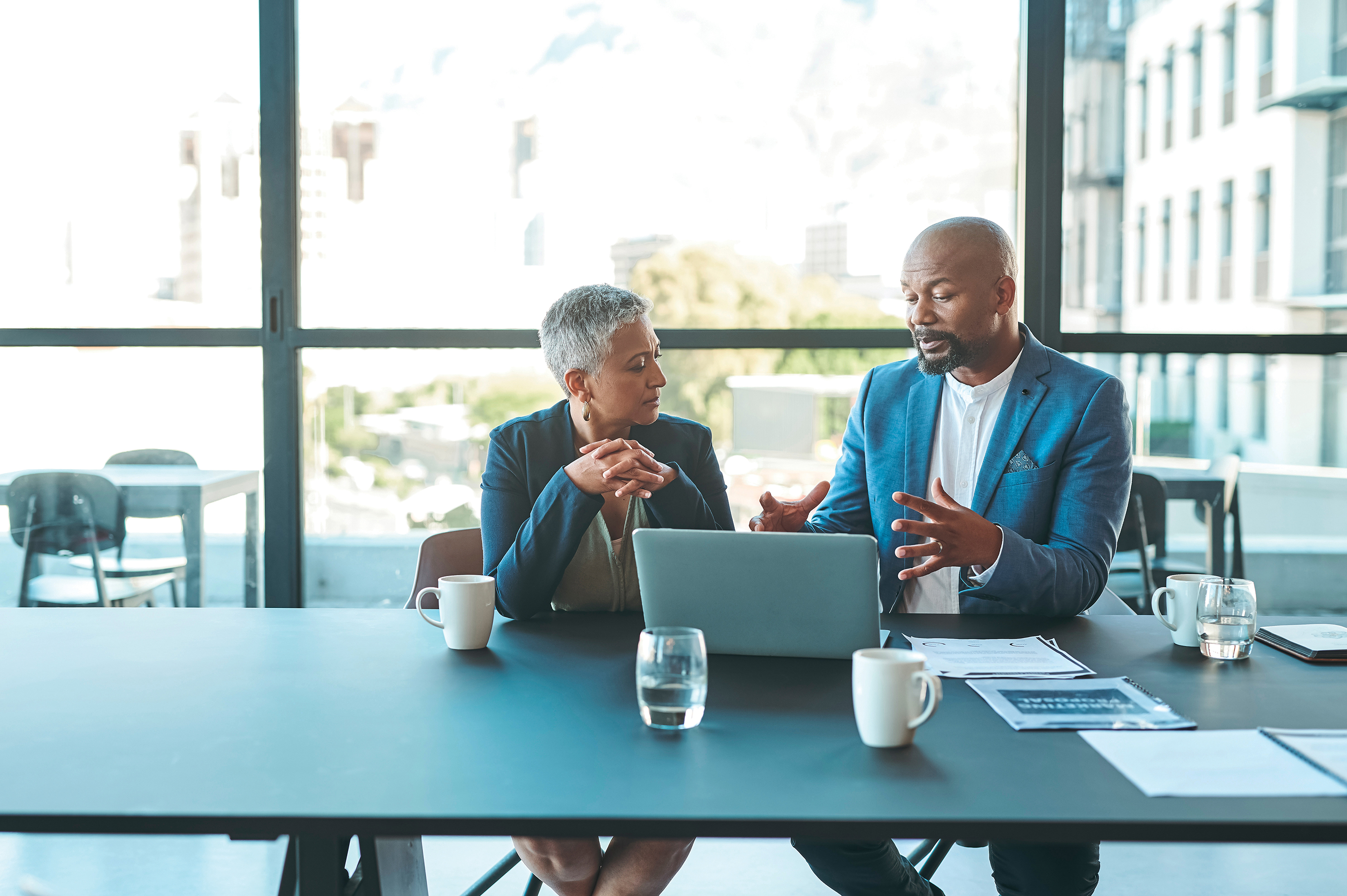 Two professionals having a meeting at a table in an office and looking at a laptop.