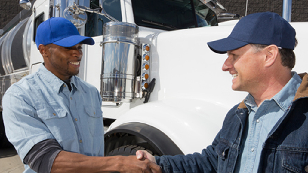 Two truck drivers wearing blue baseball caps shaking hands, white semi truck in background
