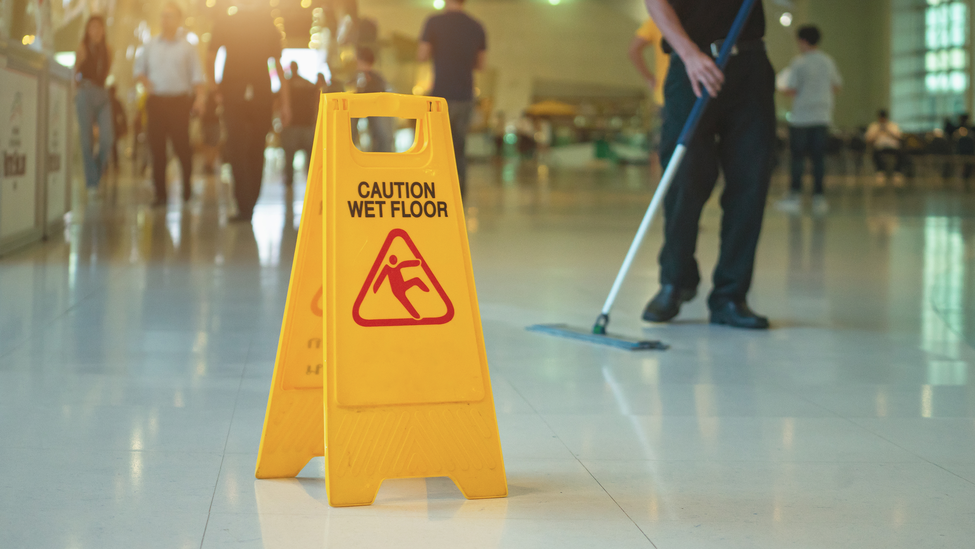 A yellow “wet floor” cone stands on shiny white tiles while people walk swiftly by. 