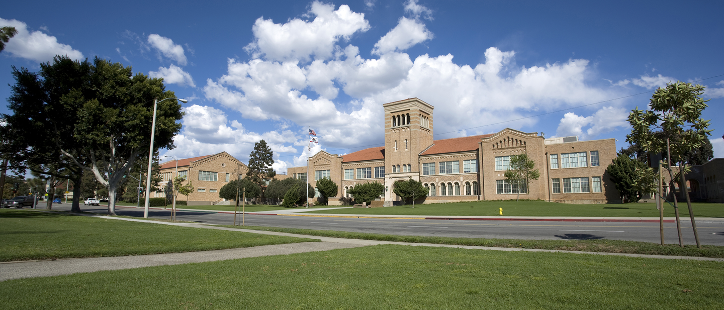 A light-colored brick school stands tall against a wide green lawn and blue sky.