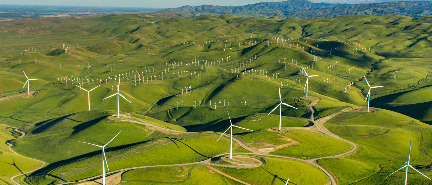 Aerial view of 10 wind turbines strewn across a green, hilly landscape.