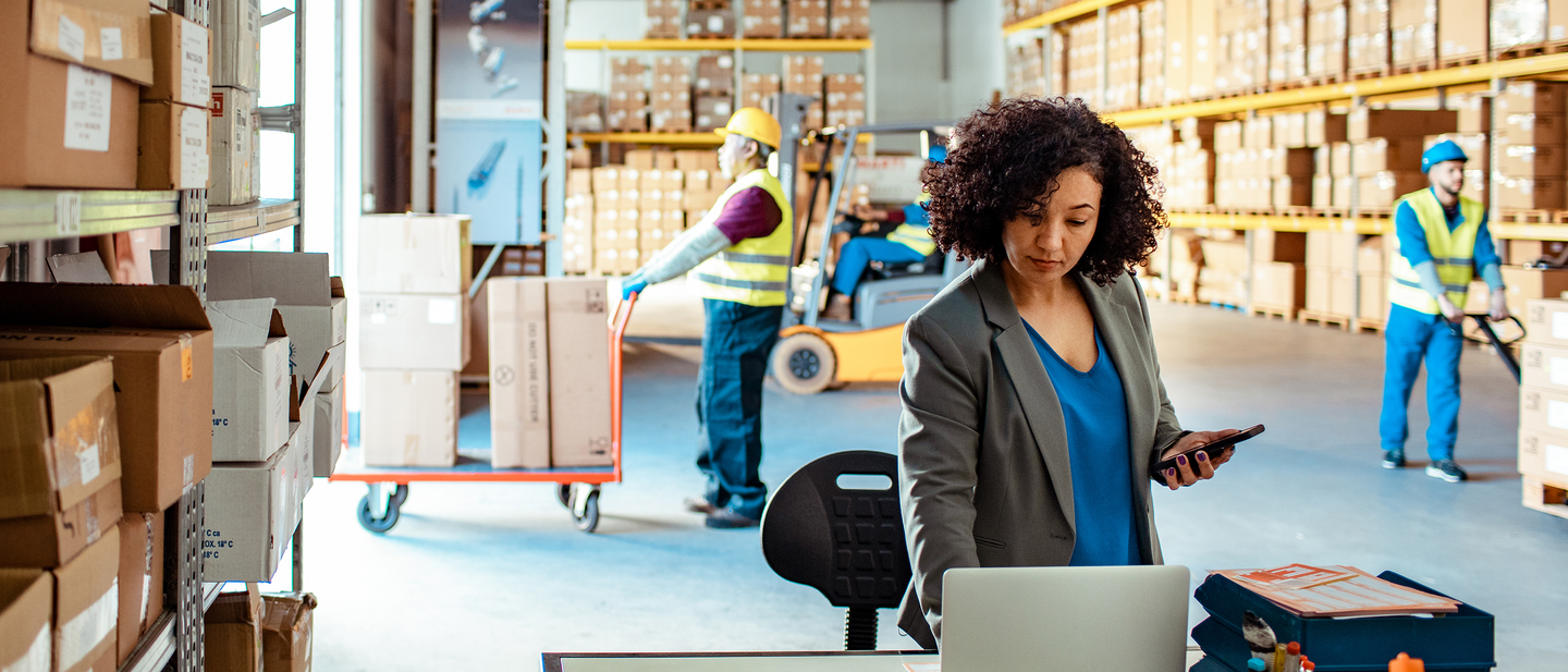 A woman in a warehouse full of boxes with men working in the background.