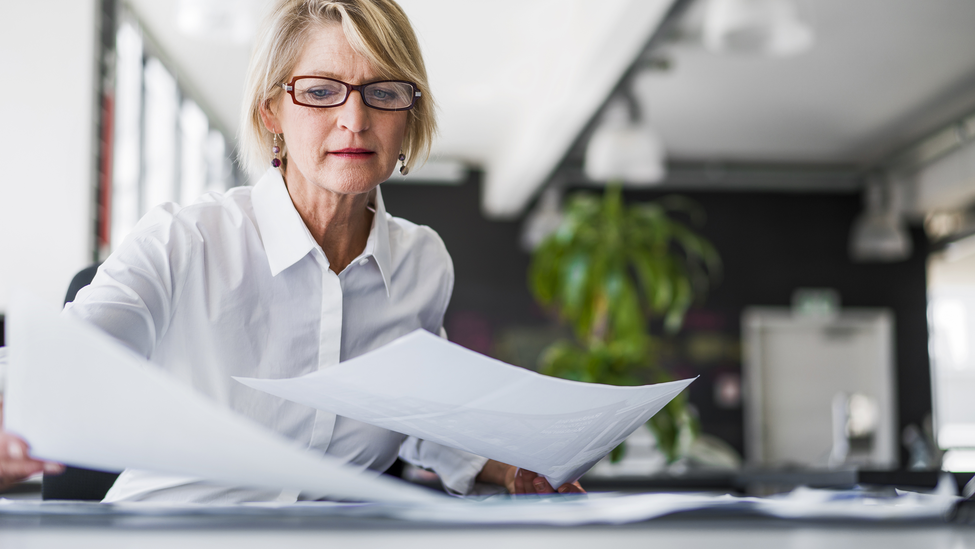 A photo of mature businesswoman examining documents at desk. Concentrated professional is analysing papers in office. Executive is in formals.; Businesswoman examining documents at desk