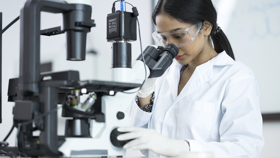 Asian Laboratory technician looking through microscope while examining genomic sample during research in laboratory.