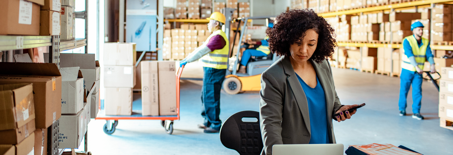 A warehouse vendor working on a laptop with warehouse workers moving product in the background.