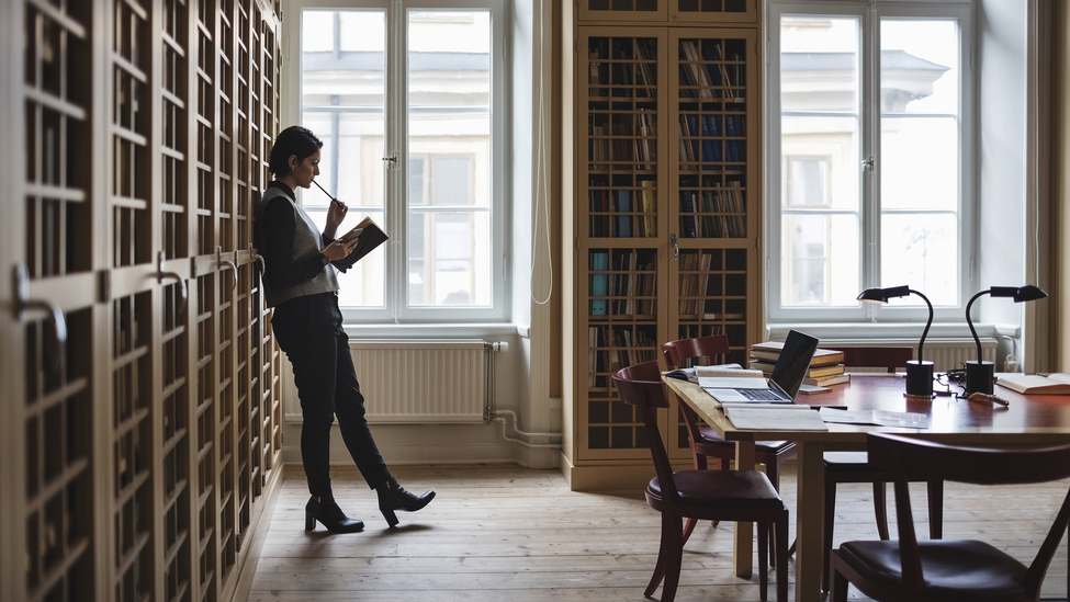 Thoughtful lawyer holding book while leaning on shelf in library