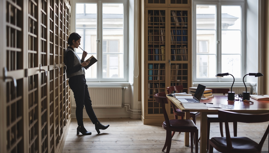 Thoughtful lawyer holding book while leaning on shelf in library