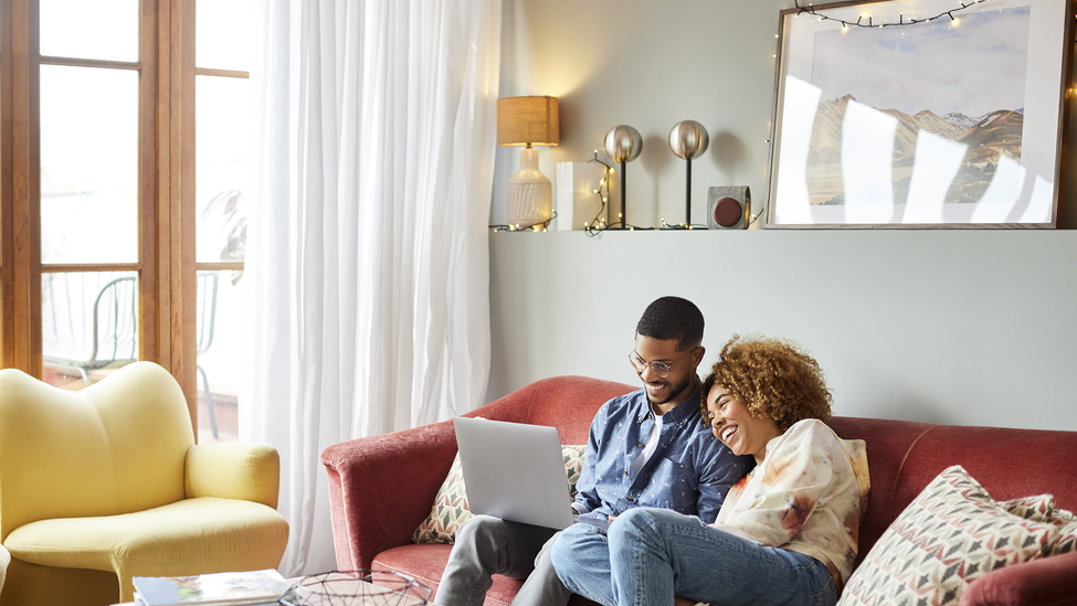 Smiling couple on a living room couch looking at home insurance information on a laptop. 