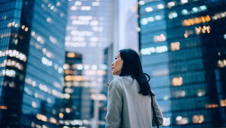 Confident and professional young Asian businesswoman looking up while standing against contemporary corporate skyscrapers with illuminated facade in financial district in the evening.