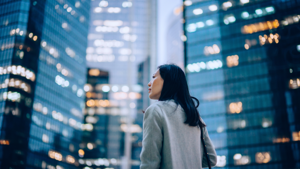 Confident and professional young Asian businesswoman looking up while standing against contemporary corporate skyscrapers with illuminated facade in financial district in the evening.