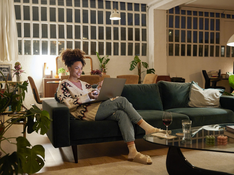 Woman on laptop snacking and relaxing in plant filled living room.
