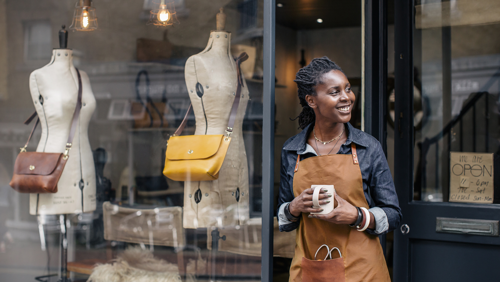 A female entrepreneur stands outside her store, with a cup of coffee. Store sells bespoke designer handbags, hand made on site in Brighton.