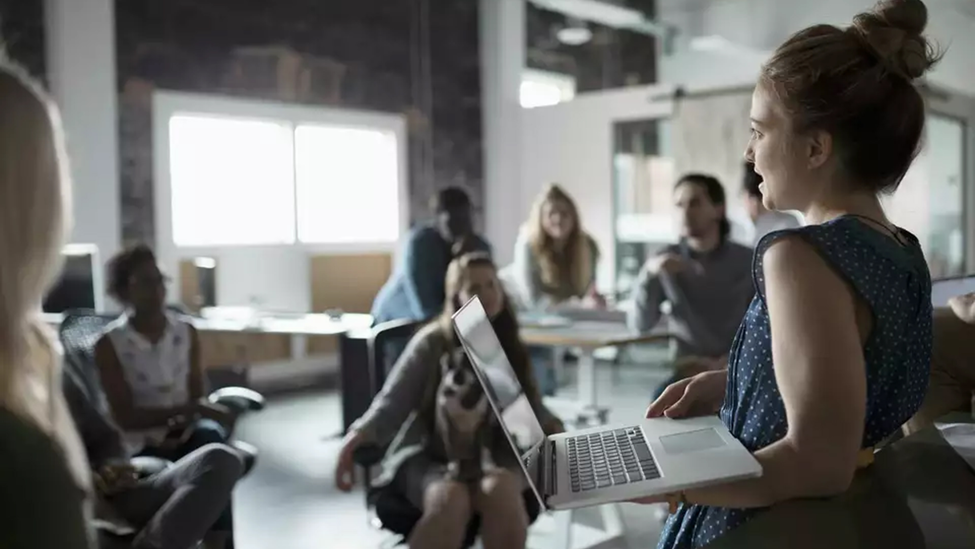 A woman holds a laptop has she speaks to a group in an office.