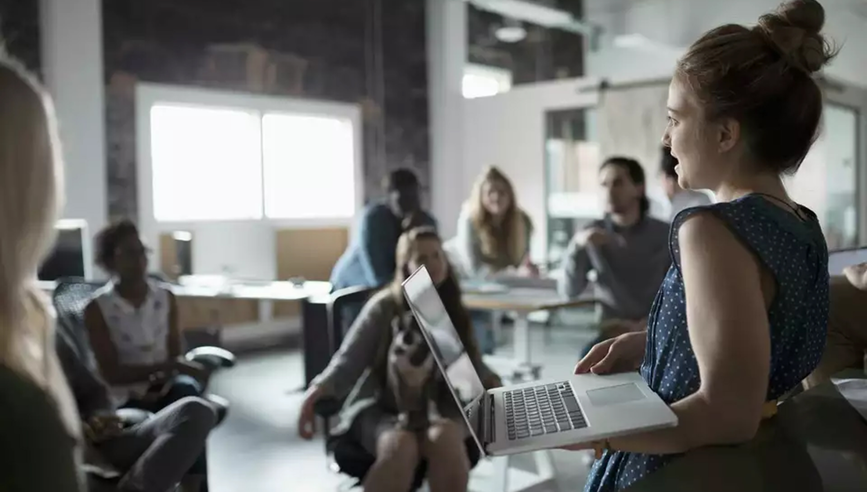 A woman holds a laptop has she speaks to a group in an office.