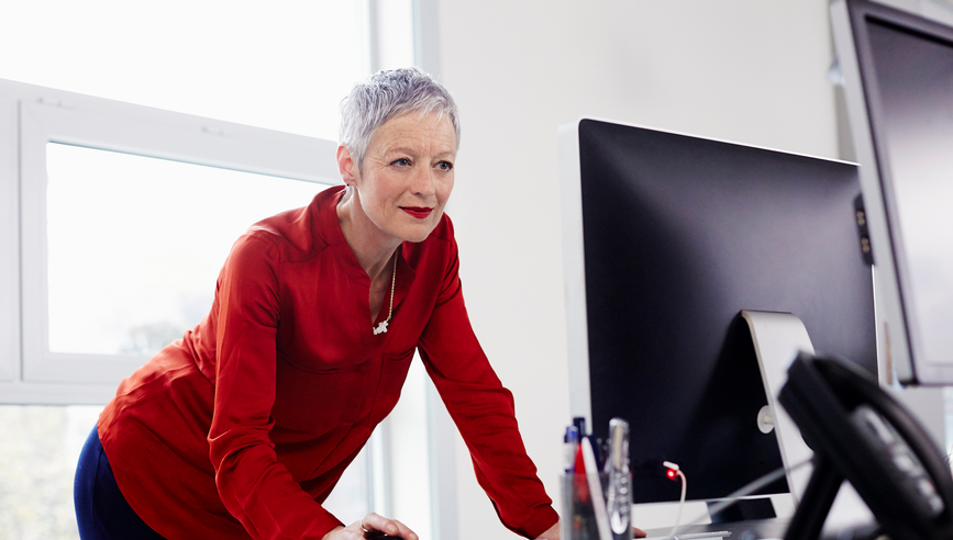 Woman leaning over desk holding mouse looking at computer monitor.