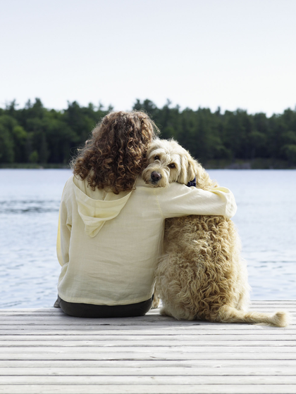 Pet owner sitting on a lake dock while hugging their dog.