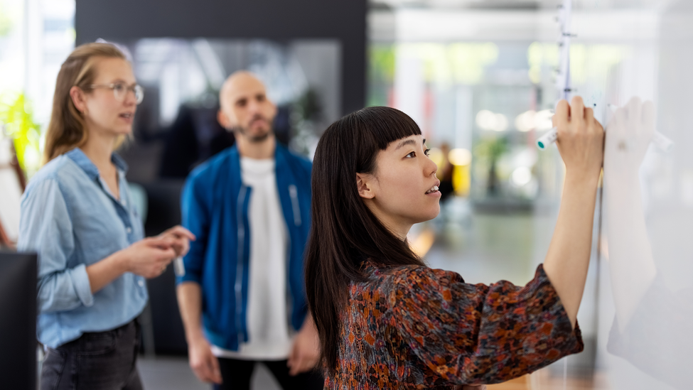 Young businesswoman explaining a business plan to colleague in office. Woman writing on the whiteboard during presentation in hybrid office space.; Businesswoman explaining new project plan to team in office