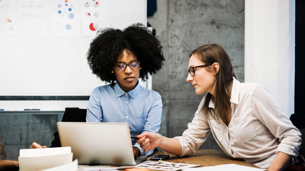 Two female coworkers look at a laptop in a business meeting in a modern office. They discuss something over papers and a laptop while one of them is pointing at the computer screen. Both are wearing glasses and pie charts are visible in the background.