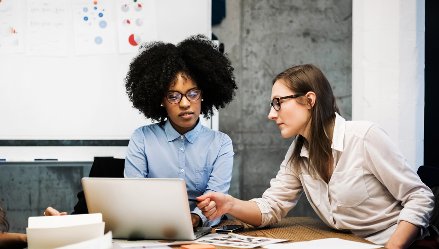 Two female coworkers look at a laptop in a business meeting in a modern office. They discuss something over papers and a laptop while one of them is pointing at the computer screen. Both are wearing glasses and pie charts are visible in the background.