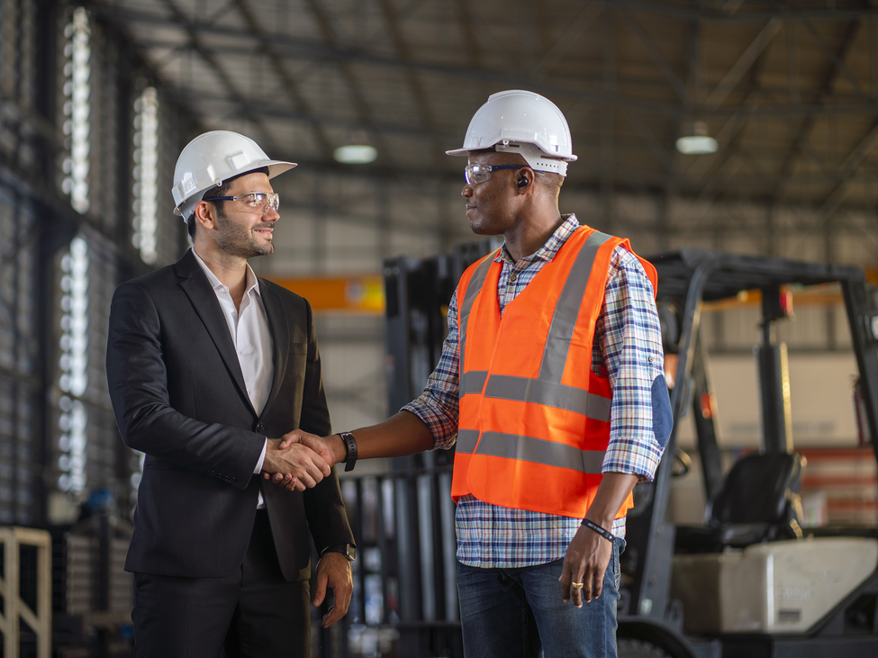 A construction worker shakes hands with a businessman in a warehouse.