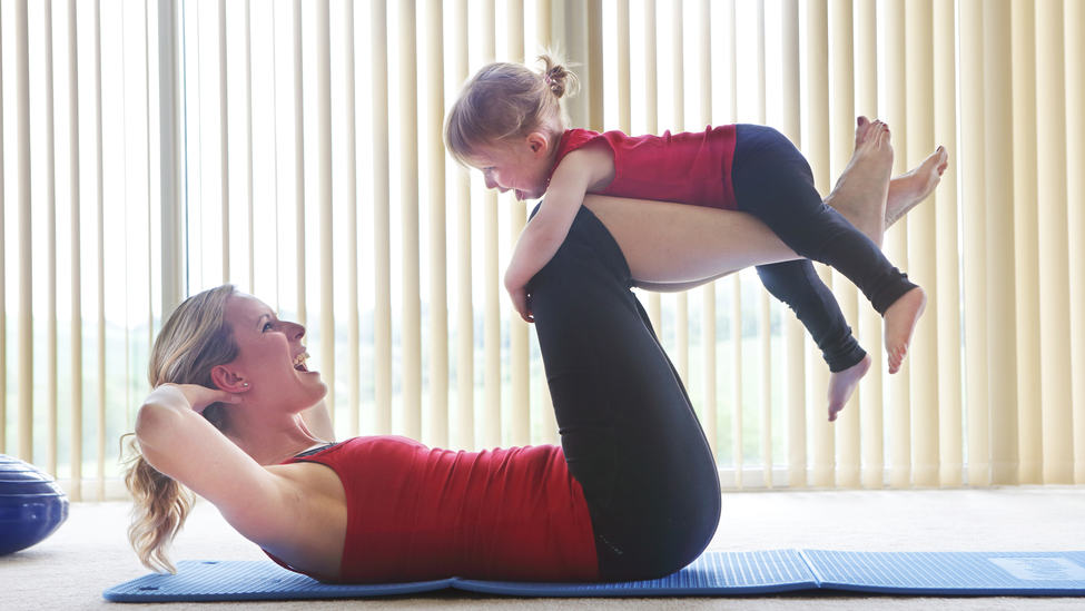 Woman lying down on a yoga mat with a child holding onto her legs.