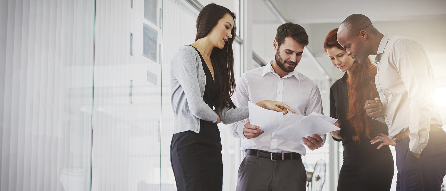 Group of four business executives reviewing documents together while standing in office.