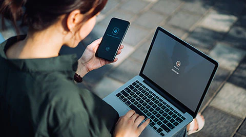 young-businesswoman-sitting-on-a-park-bench-logging-in-to-laptop-while-holding-smartphone-with-a-security-key-lock-icon-on-the-screen.jpg