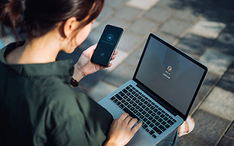 Young businesswoman sitting on a park bench, logging in to laptop while holding smartphone with a security key lock icon on the screen.