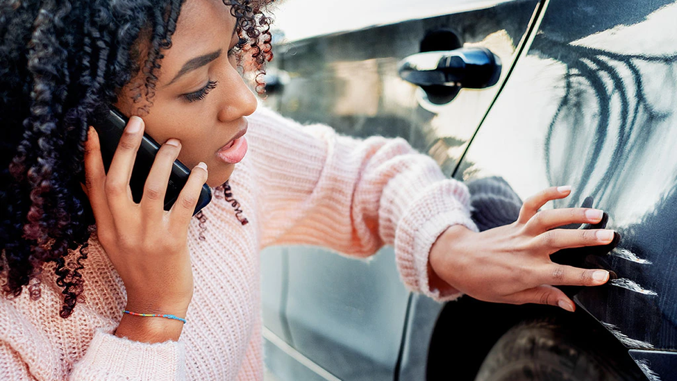 Young woman examining scratches and dents on her car, calling for help on her mobile phone.