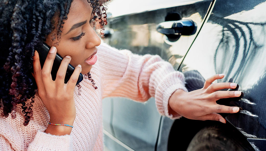 Young woman examining scratches and dents on her car, calling for help on her mobile phone.